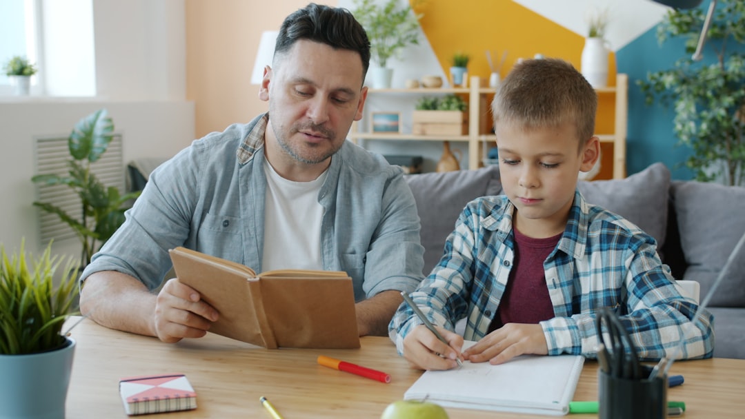 Caring dad is reading book to kid while child is drawing pictures at desk in apartment enjoying creative activity and leisure time with father. Culture and youth concept.