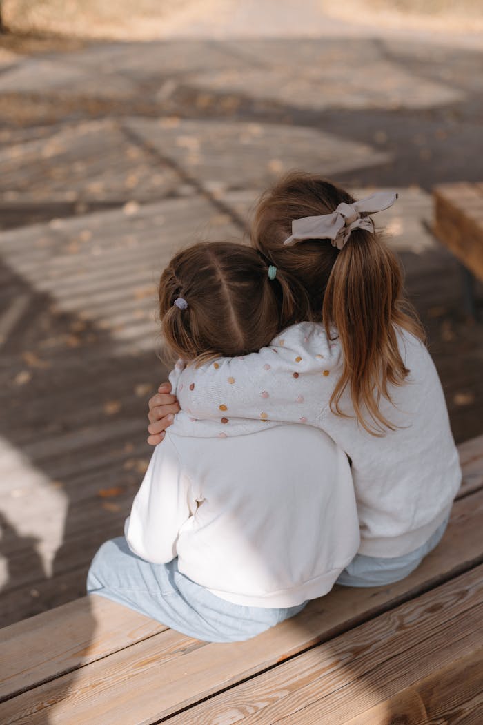 Two young girls hugging on a wooden bench, showcasing friendship and warmth in an outdoor setting.